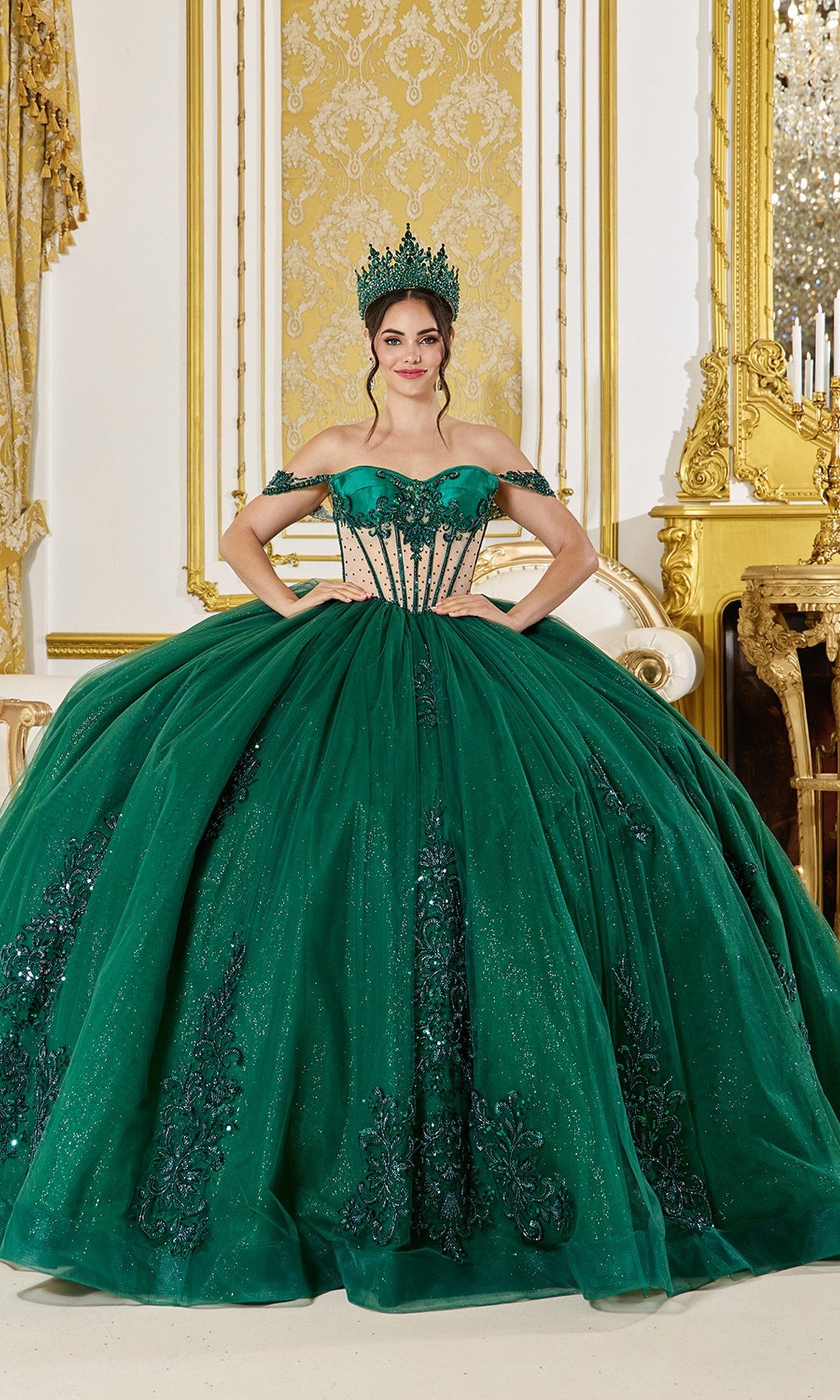A woman wears the Quinceanera Dress 5059 by Dancing Queen in hunter green, featuring a sheer corset and matching crown, standing in an ornate room with gold and cream decor.